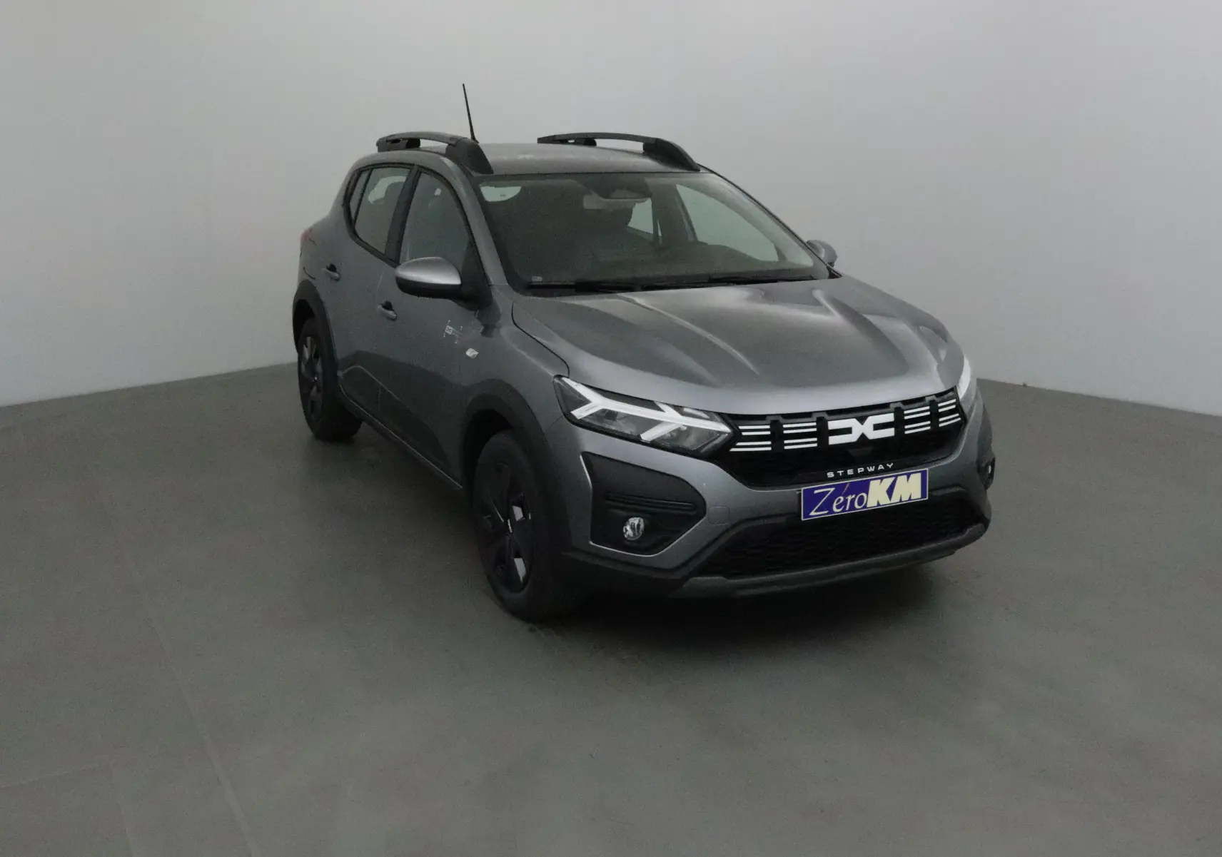 Dacia Sandero Stepway gris schiste vue 3/4 avant droit avec calandre moderne et barres de toit noires.