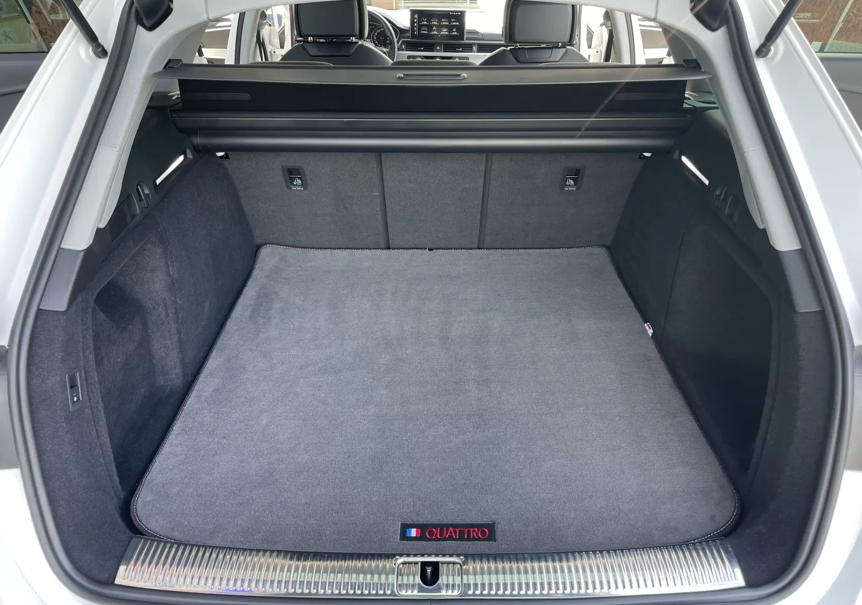 Coffre ouvert d'une Audi A4 Avant 40 TDI blanc, vue arrière, tapis gris avec logo Quattro visible au sol.