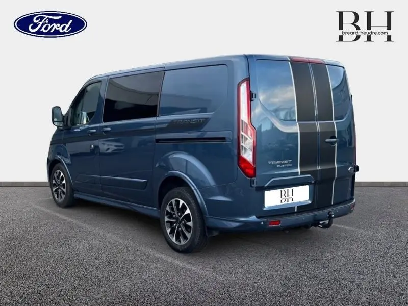 Vue 3/4 arrière droite du Ford Transit Custom Fourgon bleu azur métallisé avec bandes noires verticales et jantes alu 17 pouces.