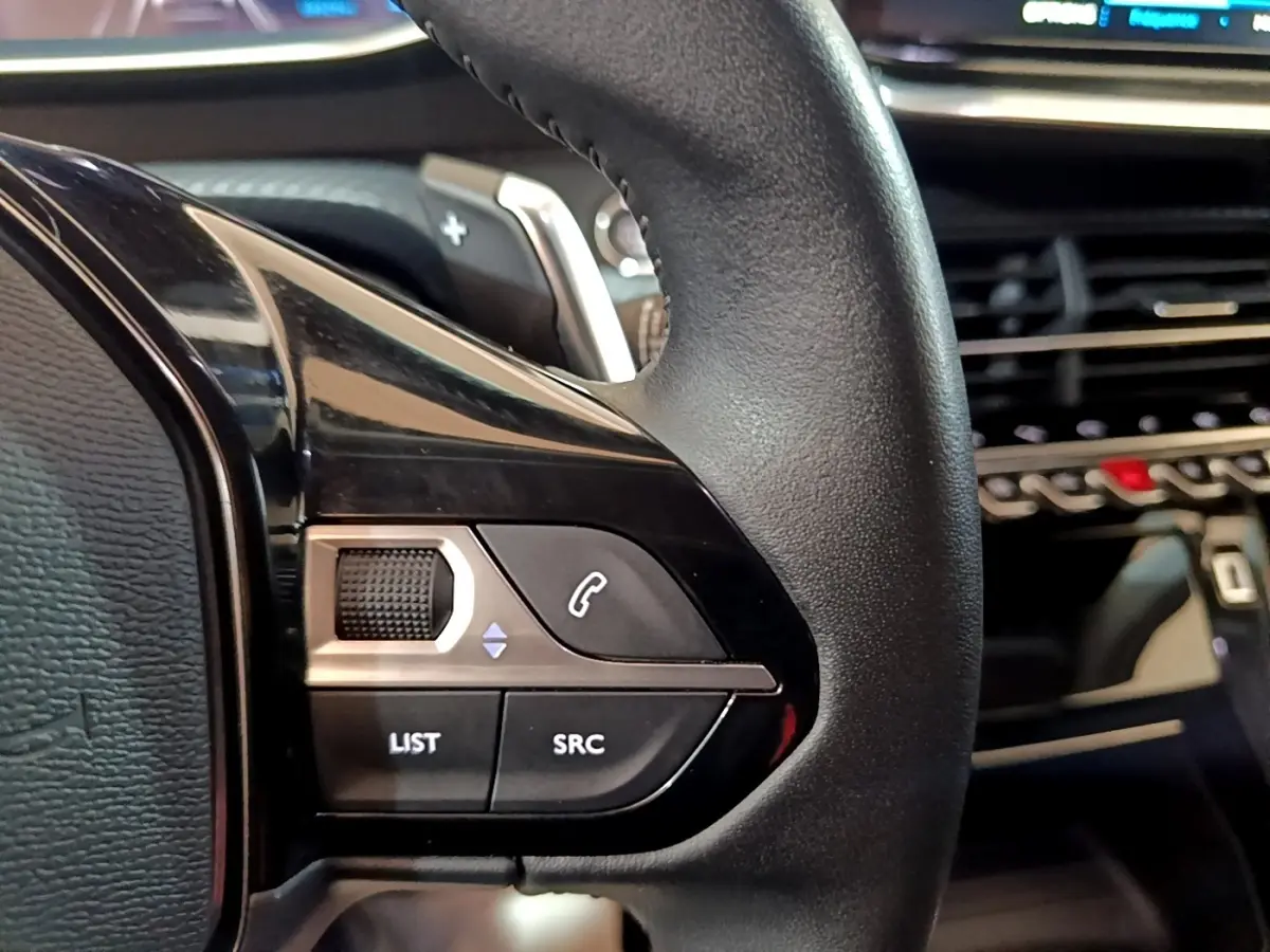 Gros plan sur les commandes gauche du volant cuir noir du Peugeot 2008, avec tableau de bord et écran tactile en arrière-plan.