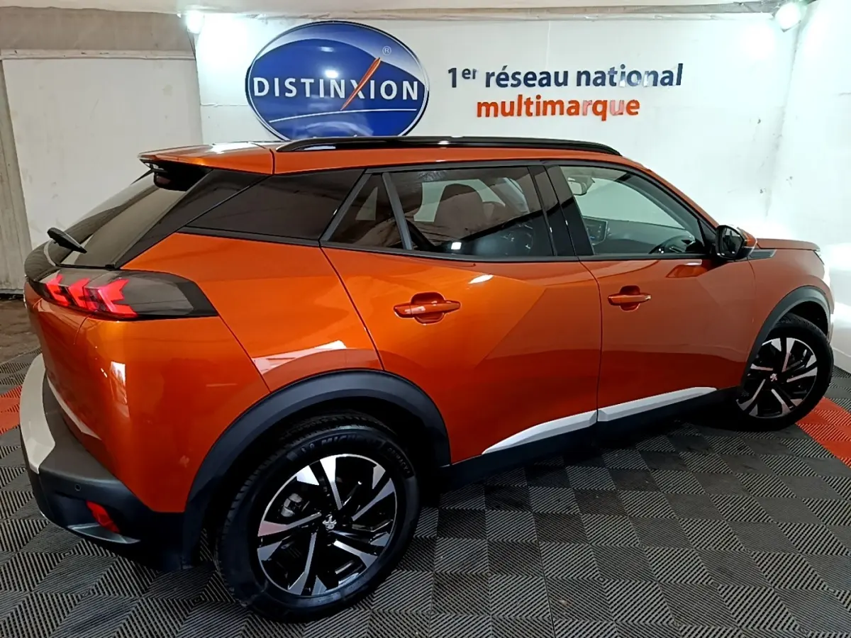 Peugeot 2008 orange vue 3/4 arrière droit, avec toit noir et jantes alliage bicolores, stationné en intérieur.