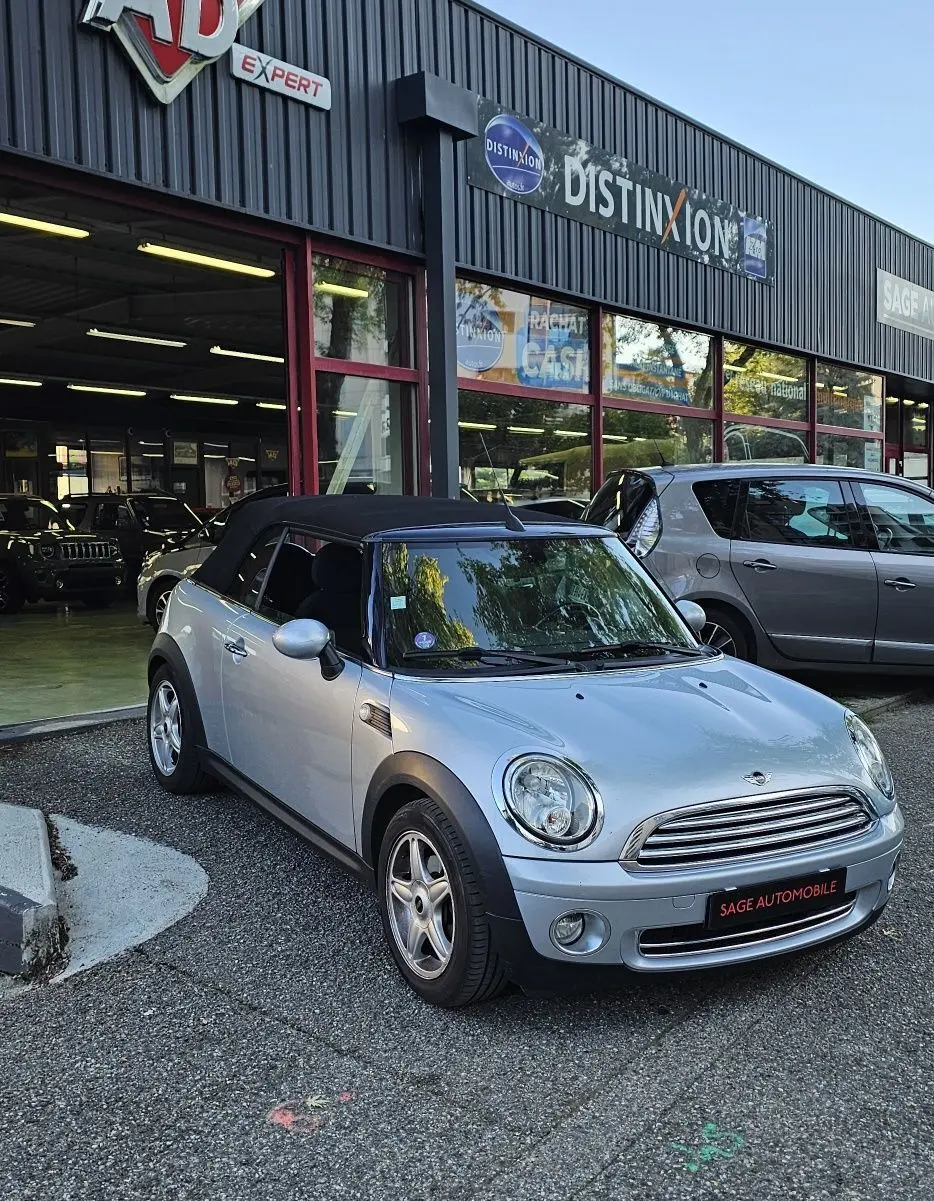MINI Cabriolet grise vue 3/4 avant droit, capote noire fermée, stationnée devant un garage automobile.