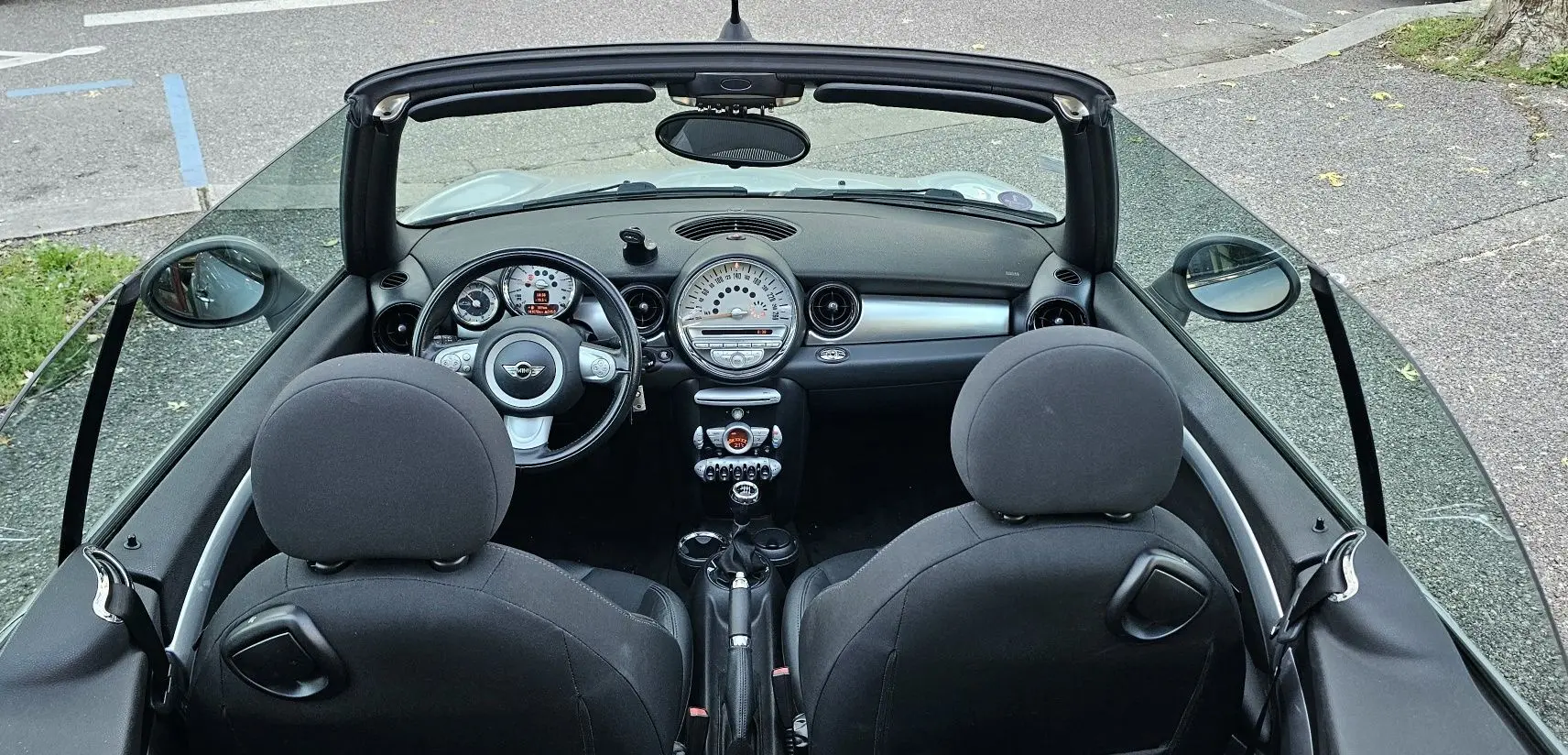 Vue intérieure de face d'une MINI Cabriolet R57 grise de 2010, montrant tableau de bord et sièges noirs ouverts.