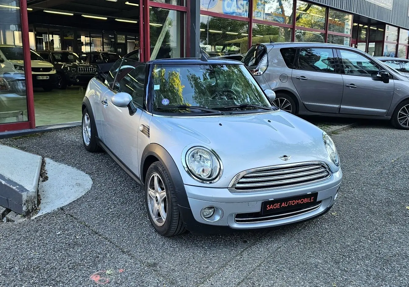 MINI Cabriolet R57 grise vue 3/4 avant droit, toit ouvert, garée devant un showroom automobile.