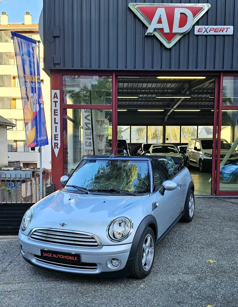 MINI Cabriolet R57 grise vue 3/4 avant droit, capote noire, stationnée devant un garage AD Expert.