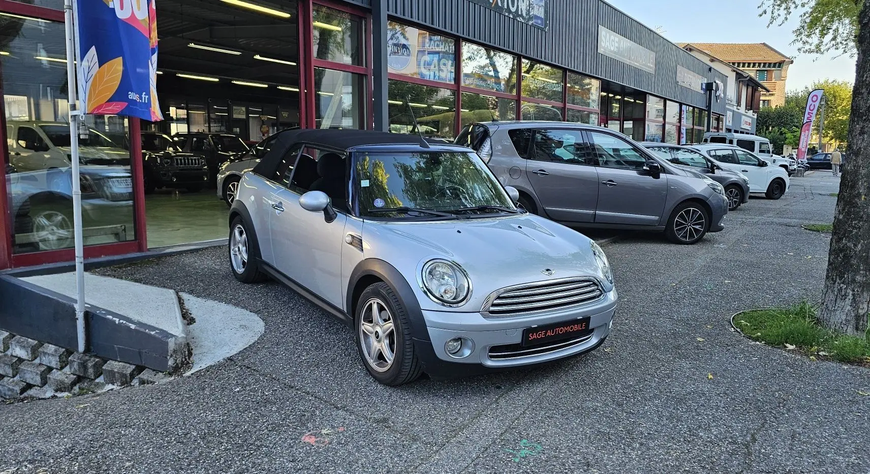 MINI Cabriolet R57 grise vue 3/4 avant droit avec capote noire, garée devant un garage automobile.
