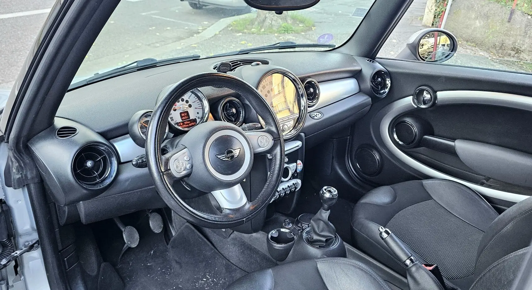 Vue intérieure côté conducteur d'une MINI Cabriolet R57 grise de 2010, avec volant cuir, tableau de bord rond et levier de vitesse manuel.