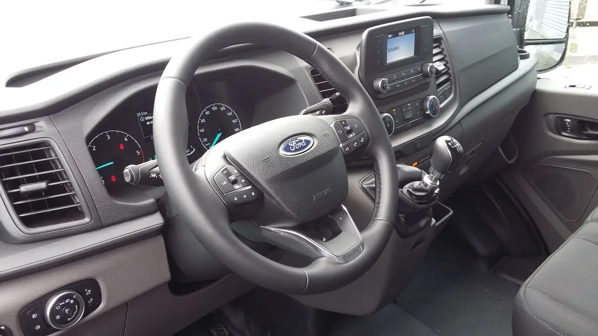 Vue intérieure côté conducteur du Ford Transit Fourgon blanc, avec volant multifonction et levier de vitesses manuel visible.