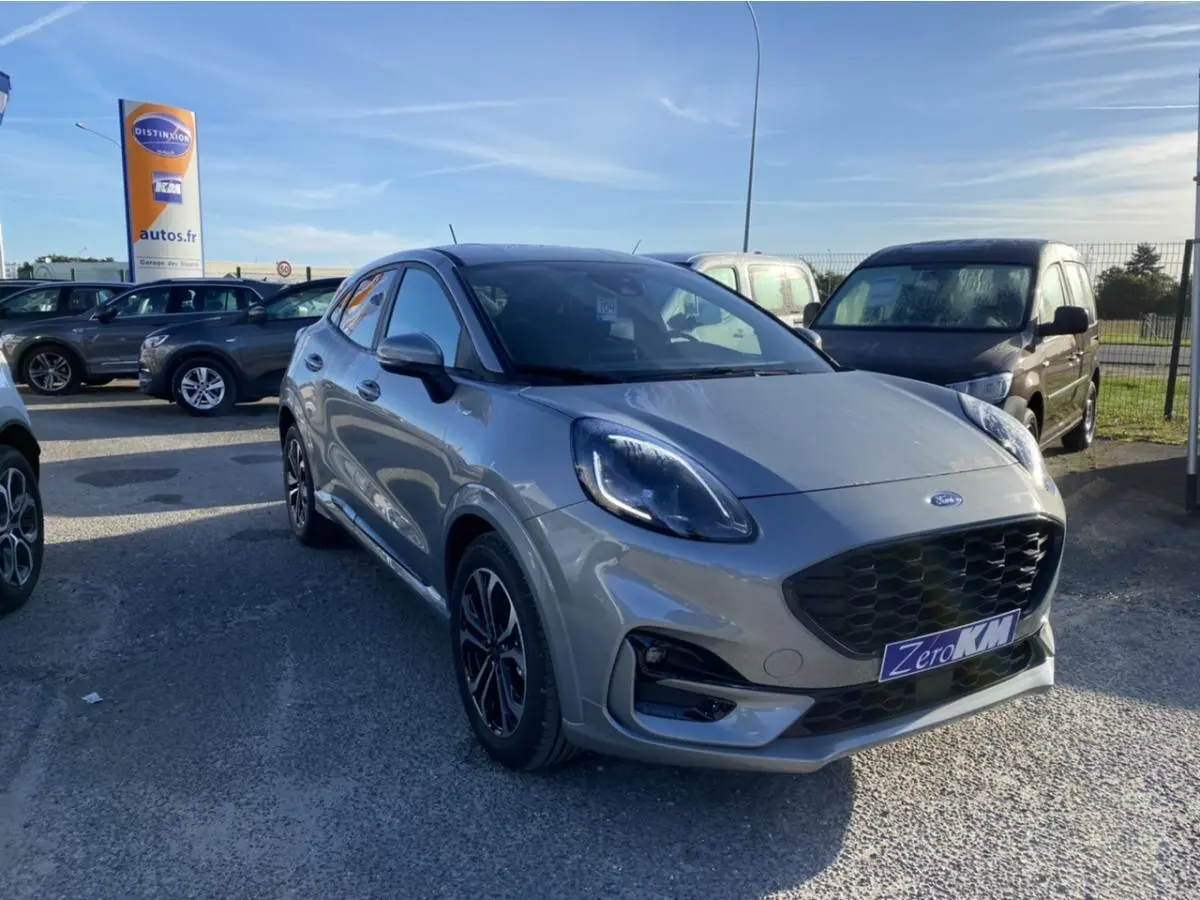 Ford Puma gris en 3/4 avant droit sur parking, avec calandre noire et jantes classiques visibles.
