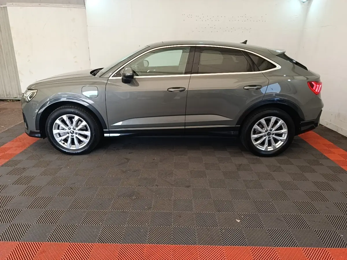 Vue de profil côté gauche d'un Audi Q3 Sportback hybride rechargeable gris métallisé avec jantes alliage et feux arrière allumés.