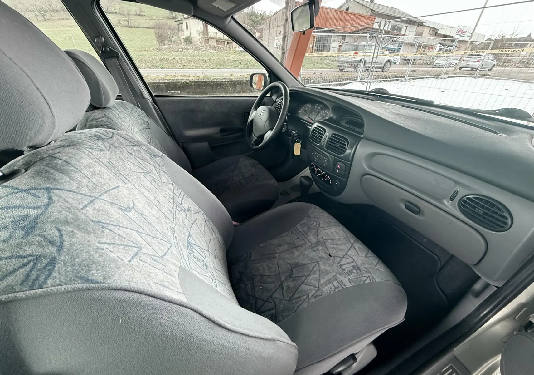 Vue intérieure côté conducteur de la Renault Mégane 2000, montrant les sièges en tissu gris et le tableau de bord simple.