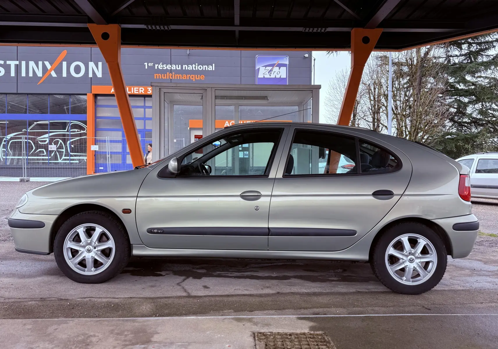Profil latéral droit d'une Renault Mégane 1.6 essence gris clair avec jantes alliage, stationnée sous abri.
