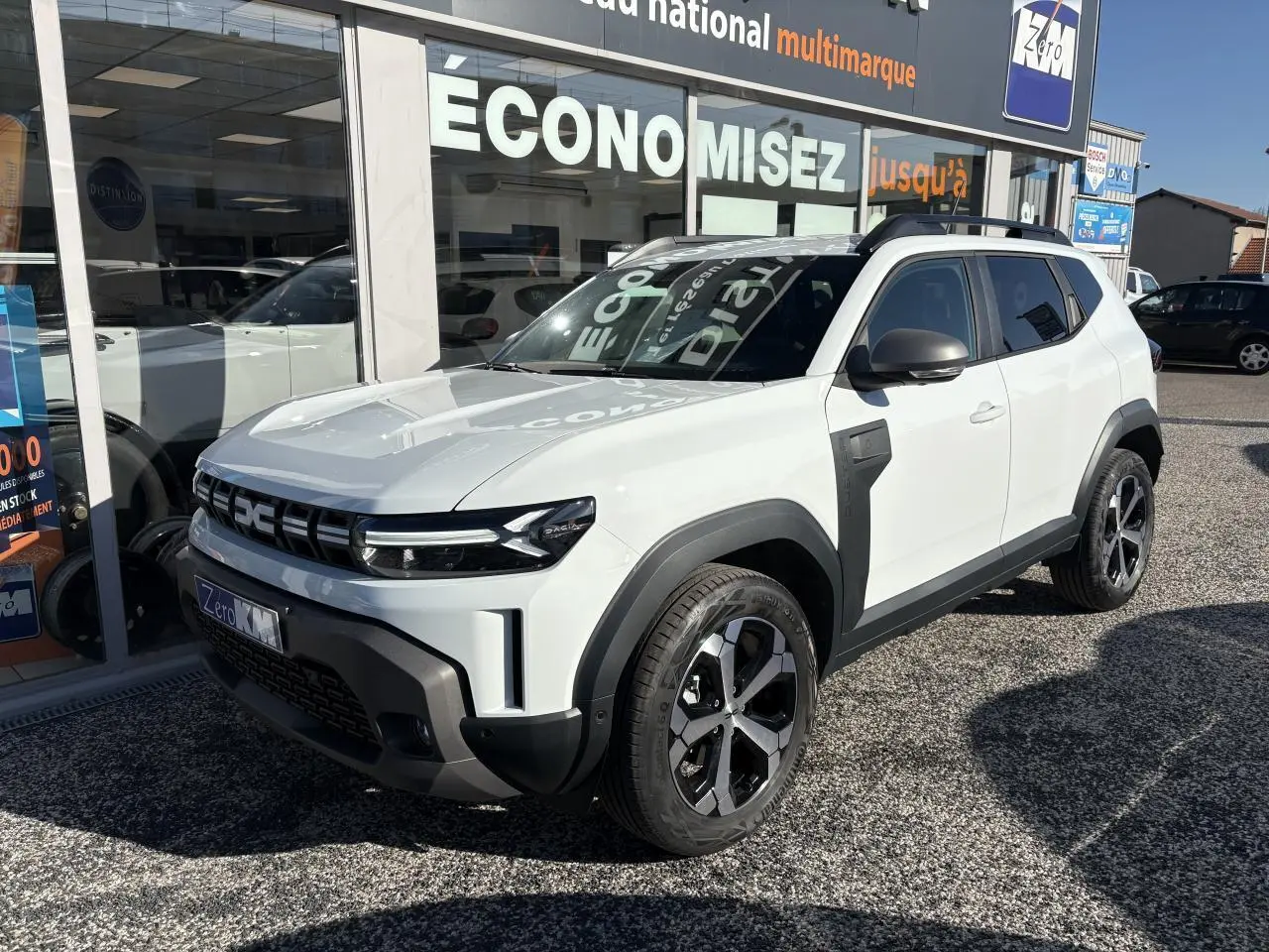 Dacia Duster blanc métallisé vu en 3/4 avant droit, avec barres de toit noires et jantes alliage distinctives.