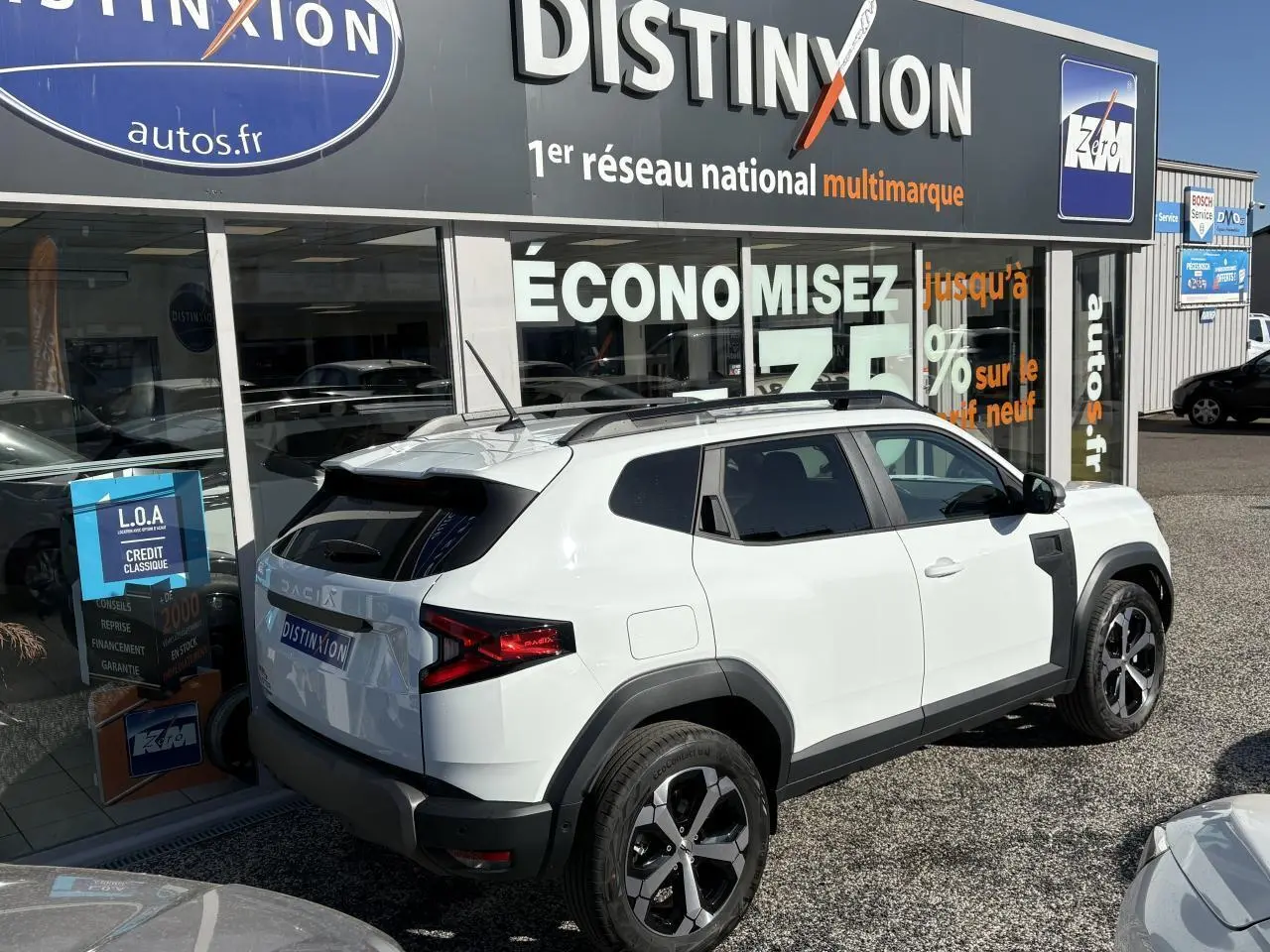 Vue 3/4 arrière droite d'un Dacia Duster 2024 blanc métallisé avec barres de toit noires et jantes alliage bicolores.