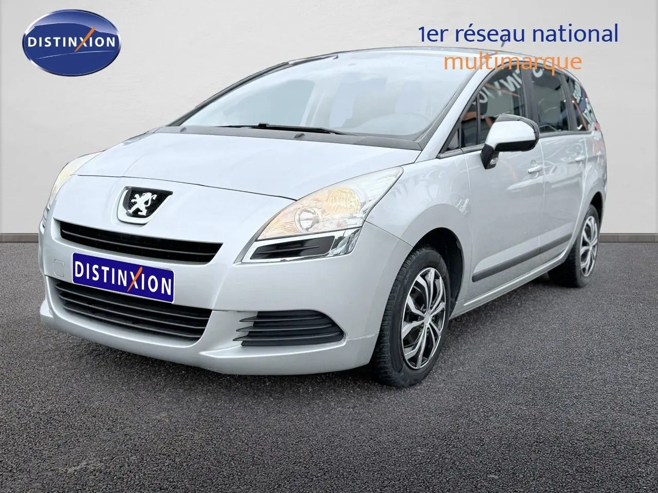 Peugeot 5008 gris vue 3/4 avant droit, montrant la calandre et les phares allumés sur fond neutre.