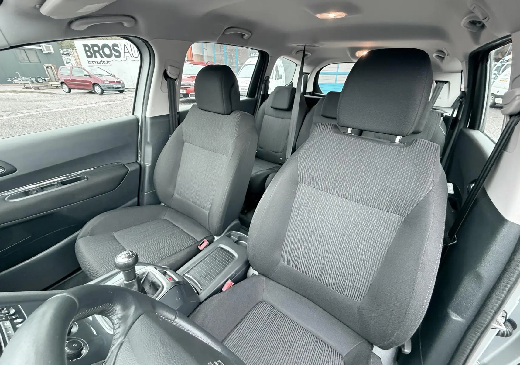 Vue intérieure du Peugeot 5008 gris, montrant les sièges avant et arrière en tissu gris avec volant cuir et levier de vitesse manuel.