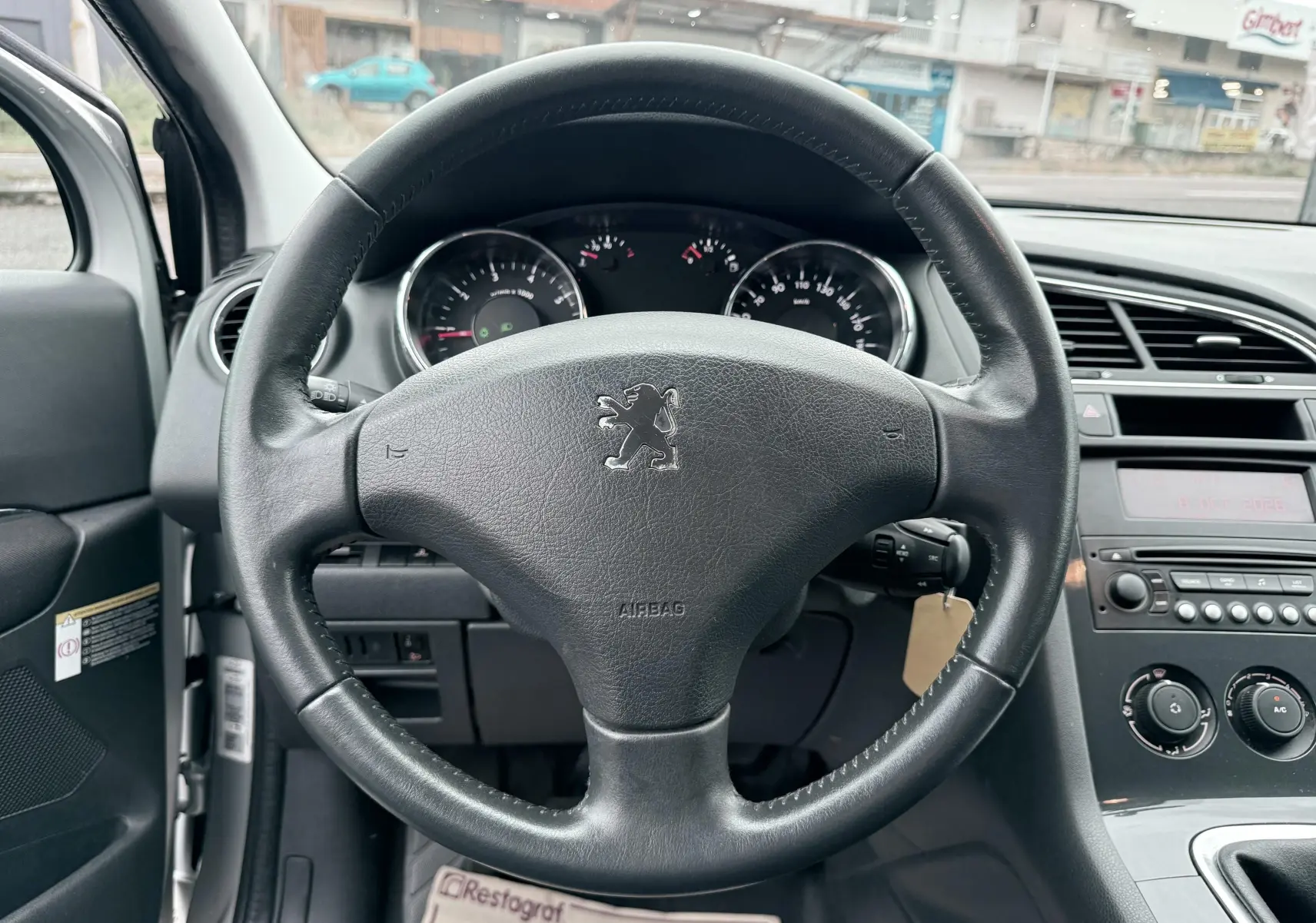 Vue intérieure centrée sur le volant en cuir noir avec logo Peugeot, tableau de bord et commandes de climatisation visibles.