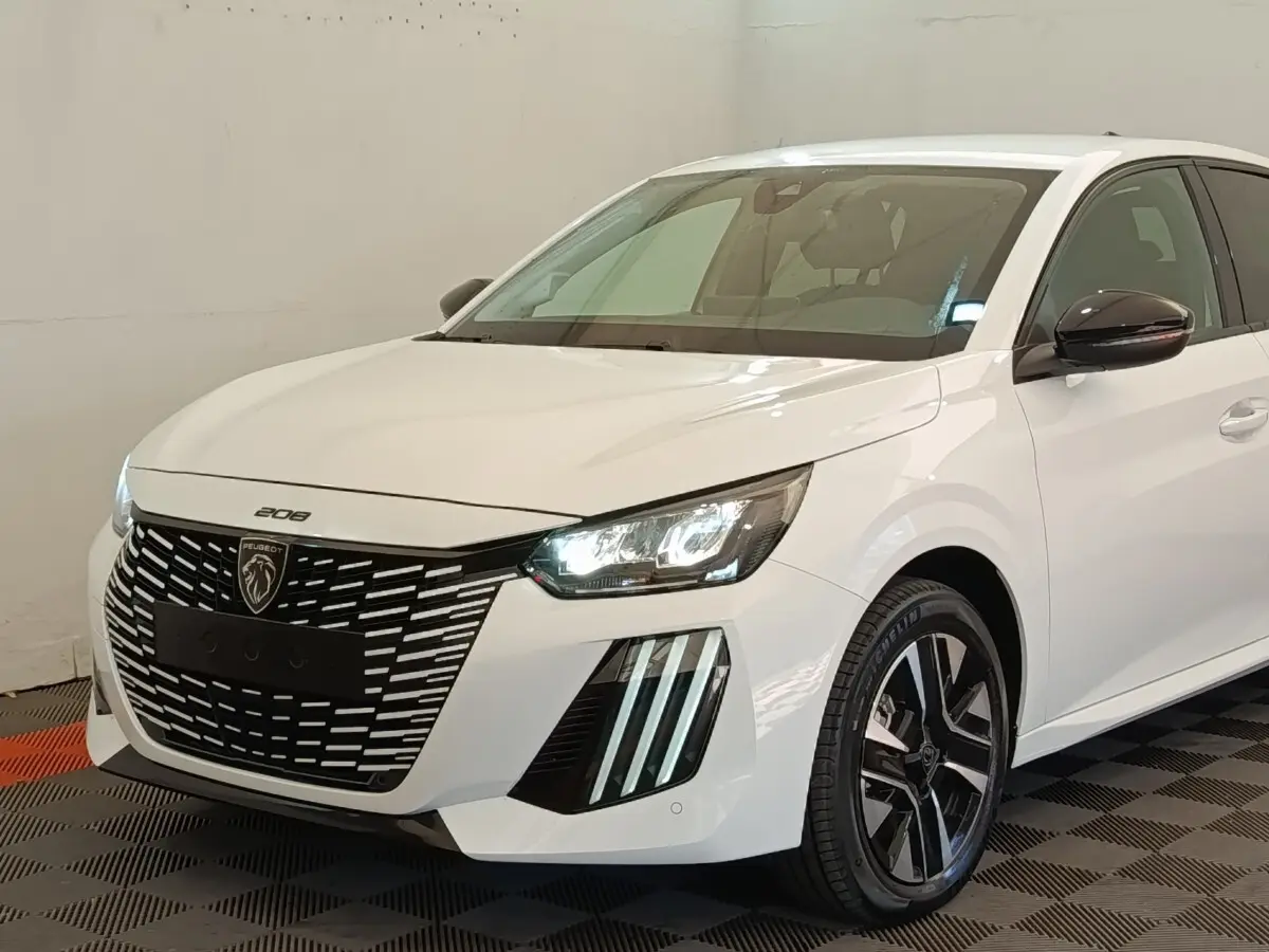 Peugeot 208 électrique blanche vue 3/4 avant droit, avec calandre noire distinctive et phares LED allumés.