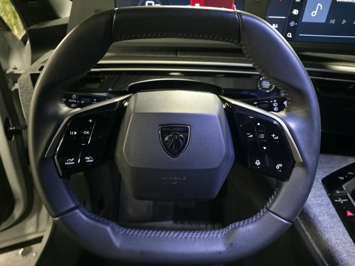 Volant compact noir avec logo Peugeot, commandes tactiles intégrées, vue frontale de l'habitacle du 5008 hybride blanc.