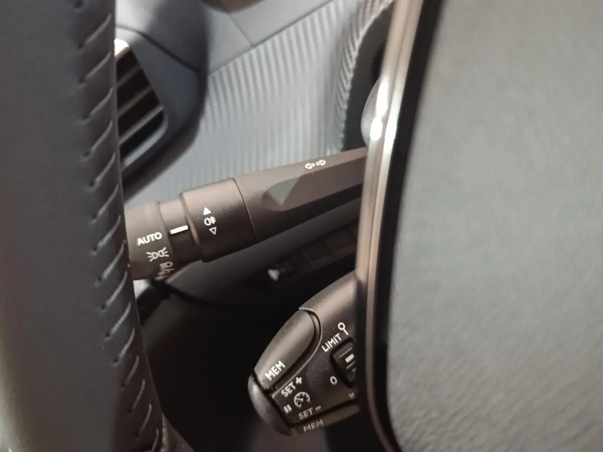 Gros plan sur les commandes au volant cuir de la Peugeot 208 électrique 2024, avec réglages du régulateur et feux automatiques.