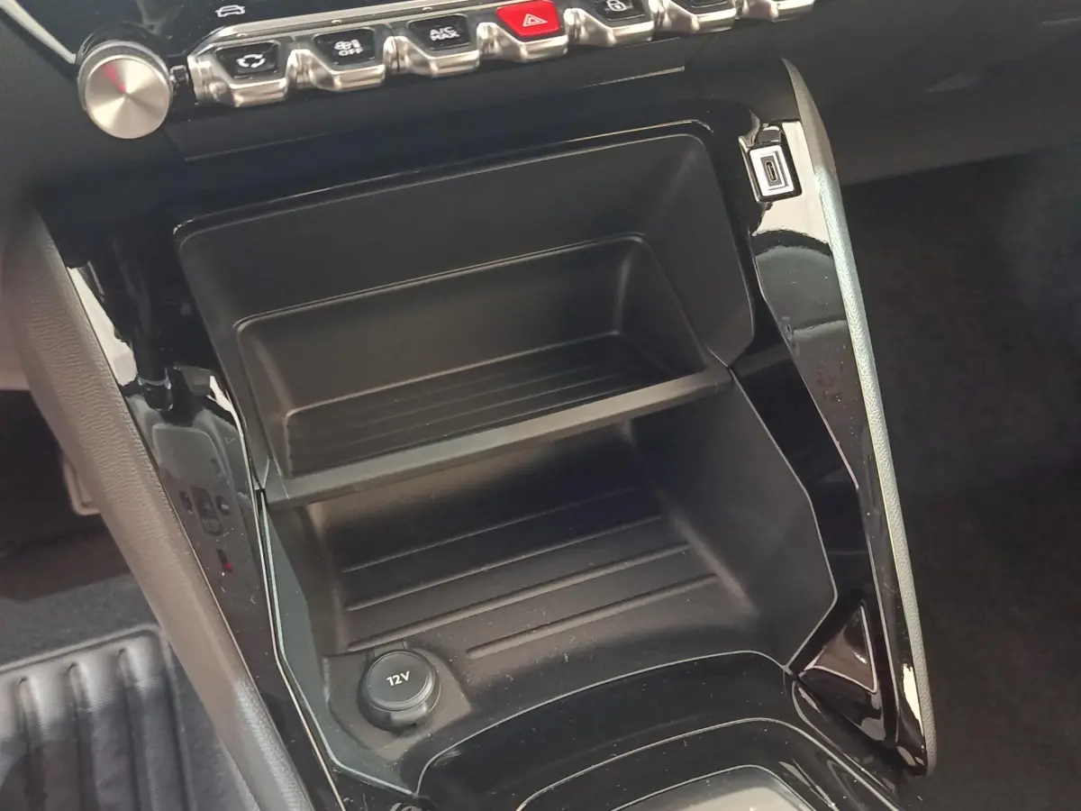 Gros plan sur la console centrale noire de la Peugeot 208 électrique 2024 avec prise 12V et rangement intégré.