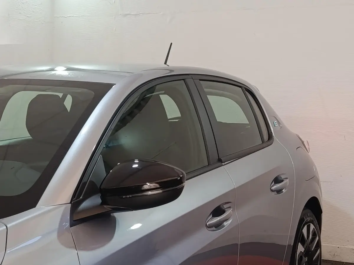 Vue côté gauche rapprochée d’une Peugeot 208 électrique gris clair avec rétroviseur noir brillant et vitres teintées.