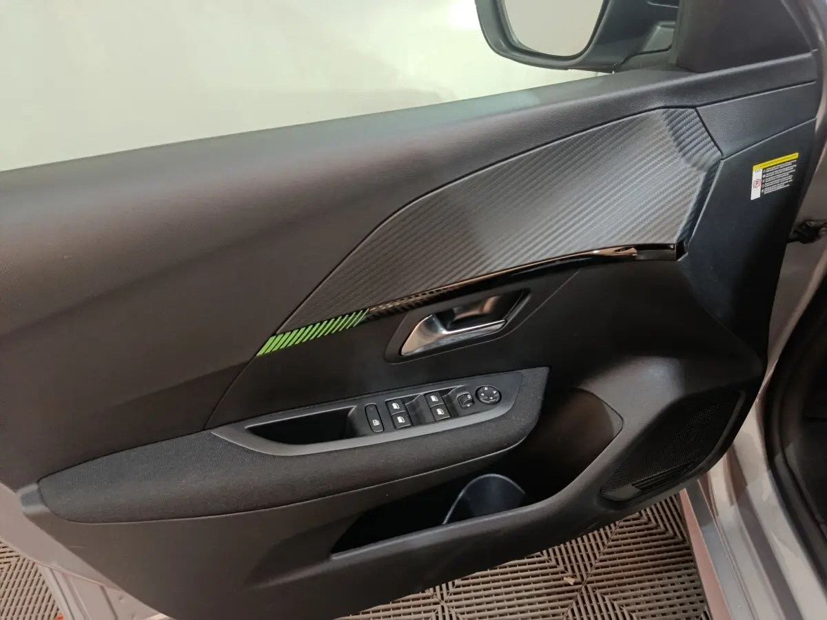 Vue intérieure côté gauche de la porte avant d’une Peugeot 208 électrique gris clair avec insert décoratif carbone et commandes fenêtres.