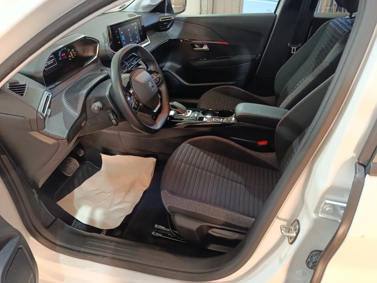 Vue intérieure côté conducteur de la Peugeot 208 électrique blanche, volant cuir multifonctions et écran tactile central visible.