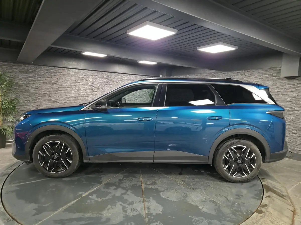 Profil droit d'un Peugeot 5008 bleu métallisé 2025 avec jantes alliage 19'' noires et barres de toit noires brillantes.