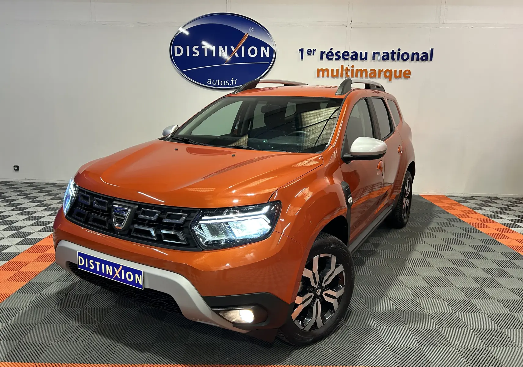 Dacia Duster orange Arizona en 3/4 avant droit, avec phares allumés et jantes alliage distinctives.