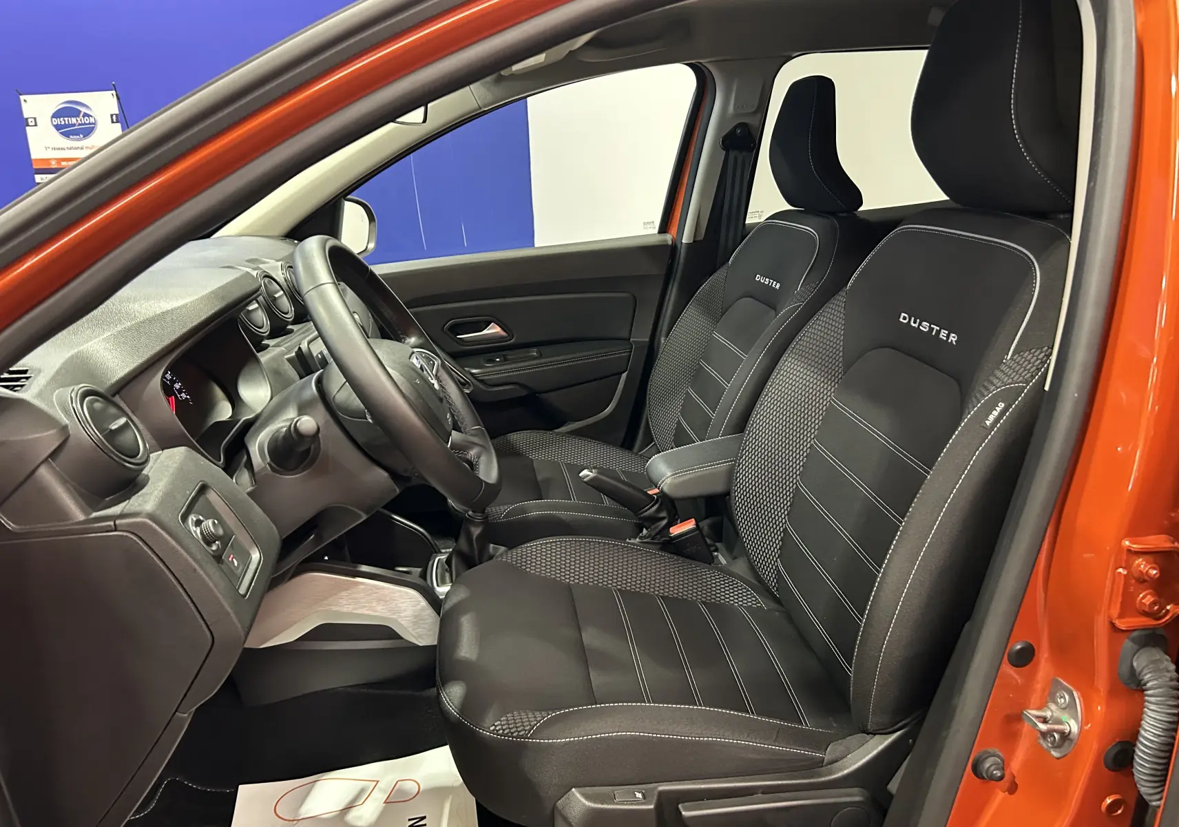Intérieur côté conducteur du Dacia Duster orange Arizona 2022, sièges noirs avec logo Duster et volant multifonction.