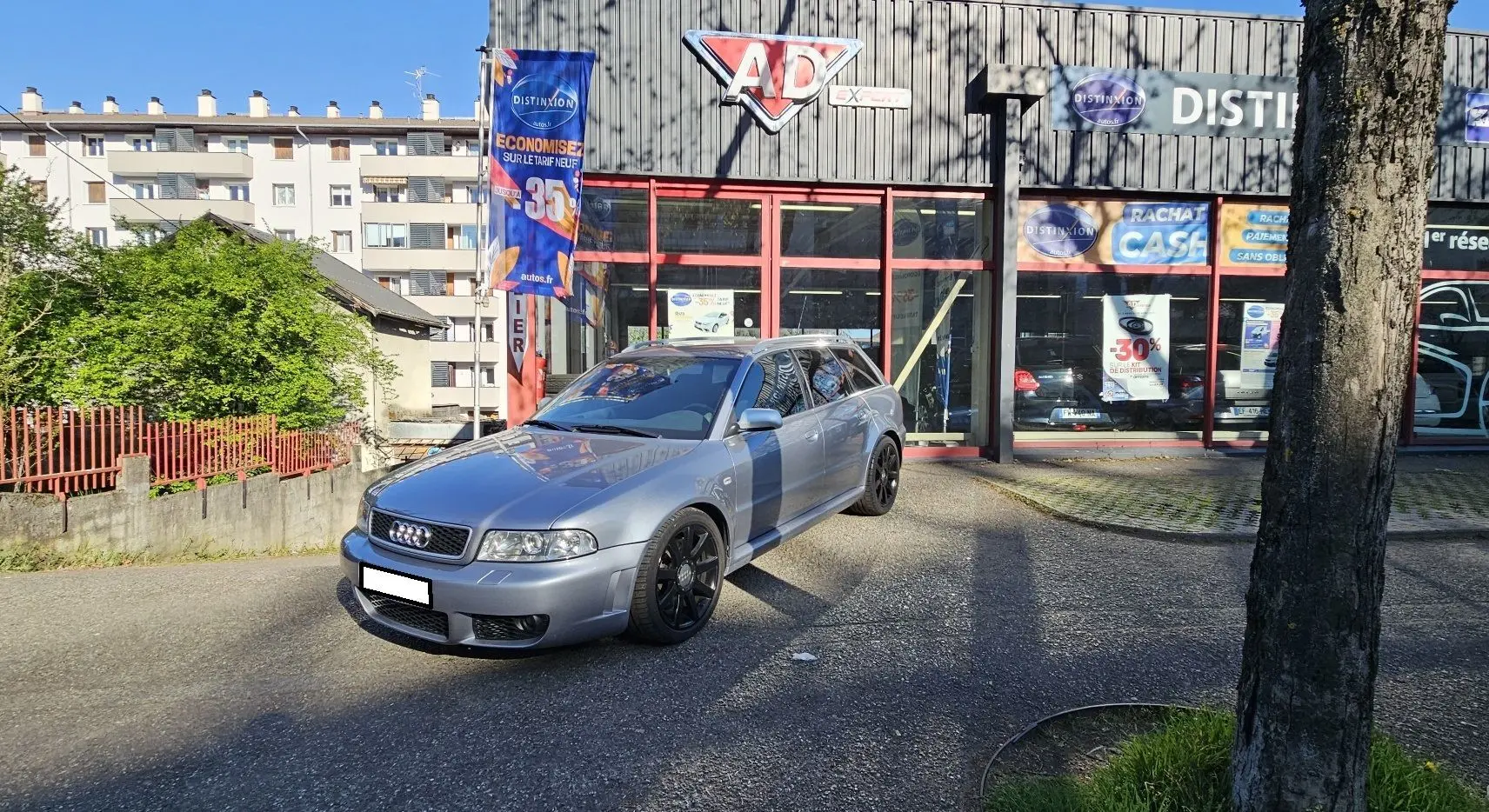 Audi RS4 Avant gris en 3/4 avant droit, avec jantes noires, stationnée devant un garage ensoleillé.