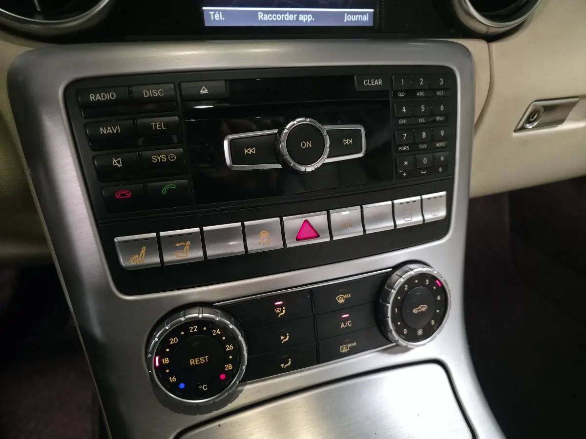 Vue rapprochée de la console centrale argentée avec commandes de climatisation et système audio d'une Mercedes SLK gris intérieur beige.