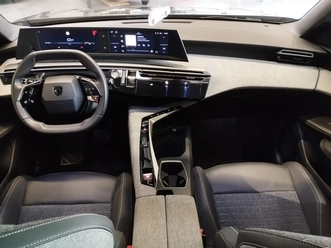 Intérieur Peugeot 5008 2025 vu de face, tableau de bord moderne avec double écran et sellerie tissu gris et cuir noir.