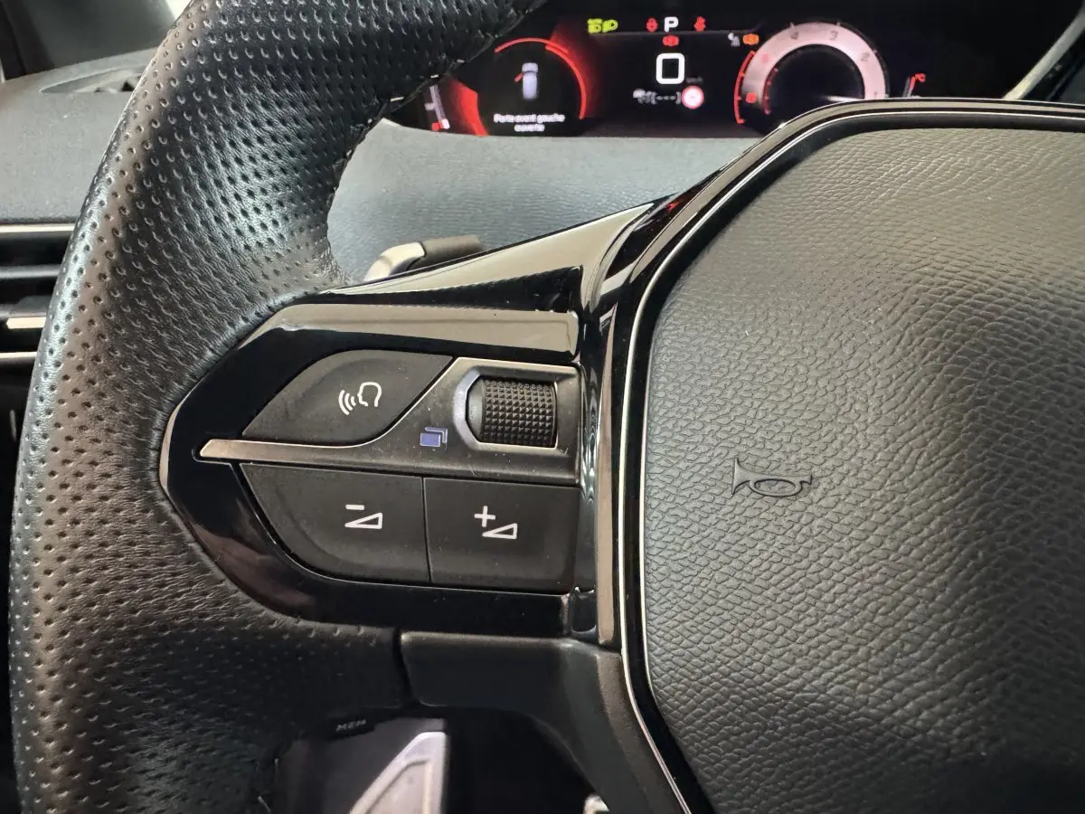 Gros plan sur le volant en cuir noir perforé du Peugeot 3008 2021, avec commandes audio et tableau de bord numérique rouge.