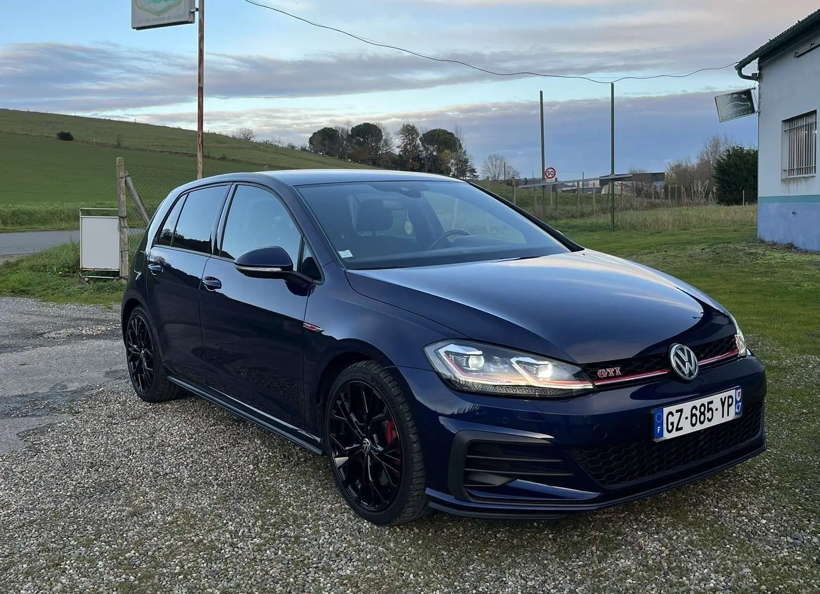 Volkswagen Golf VII GTI Performance bleu lapiz métal vue 3/4 avant droit sur parking gravier.