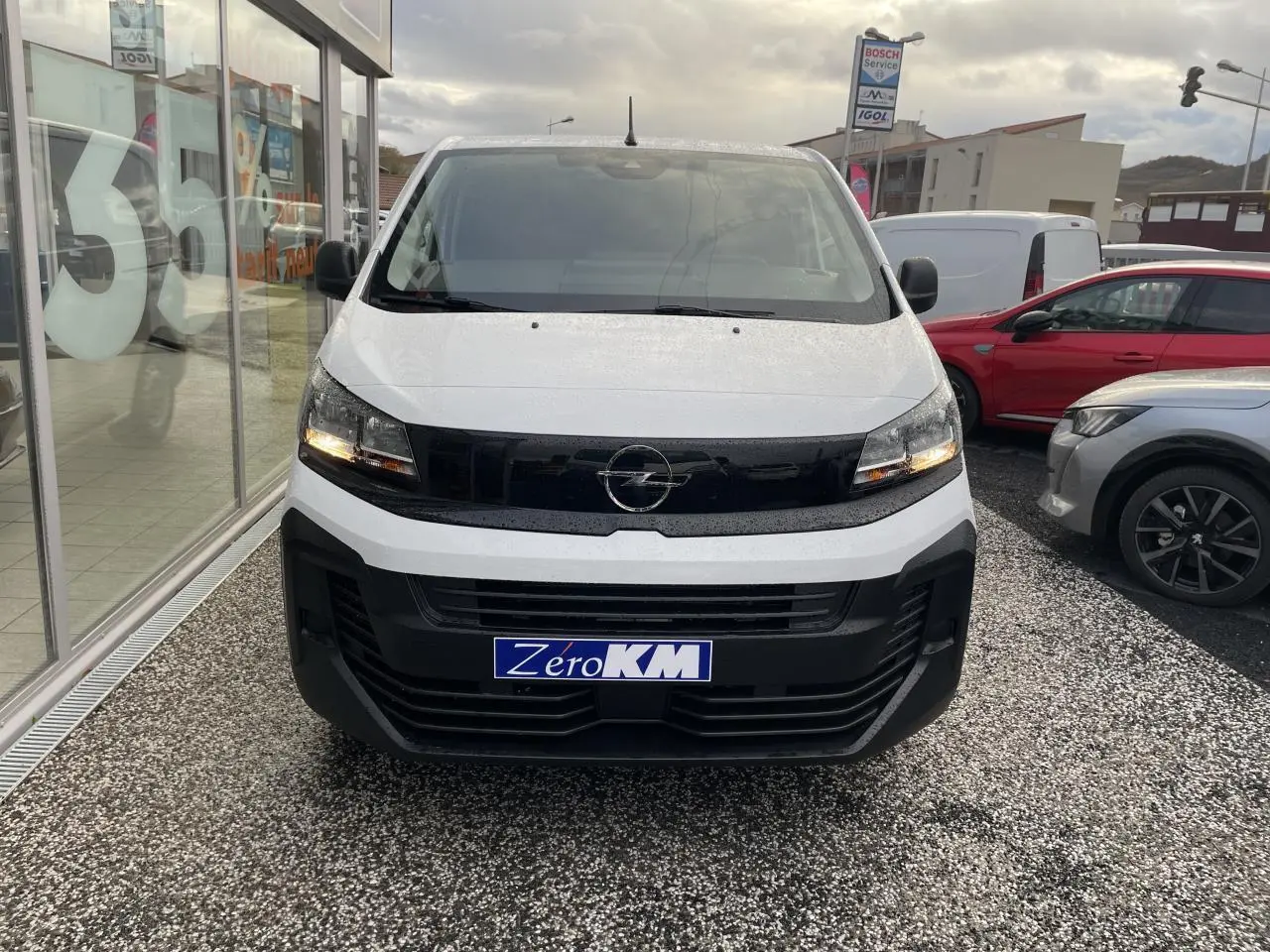 Vue frontale d'un Opel Vivaro blanc avec calandre noire et phares allumés sur parking extérieur.