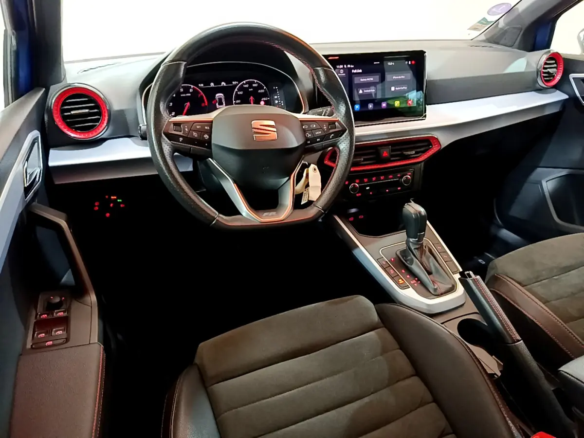 Vue intérieure avant du poste de conduite de la SEAT Arona bleu, volant cuir avec logo, écran tactile et aérateurs rouges.