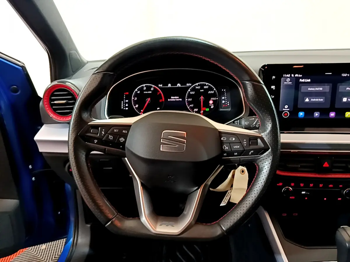 Vue intérieure centrée sur le volant cuir perforé avec surpiqûres rouges du SEAT Arona bleu 2022, tableau de bord digital et écran tactile.