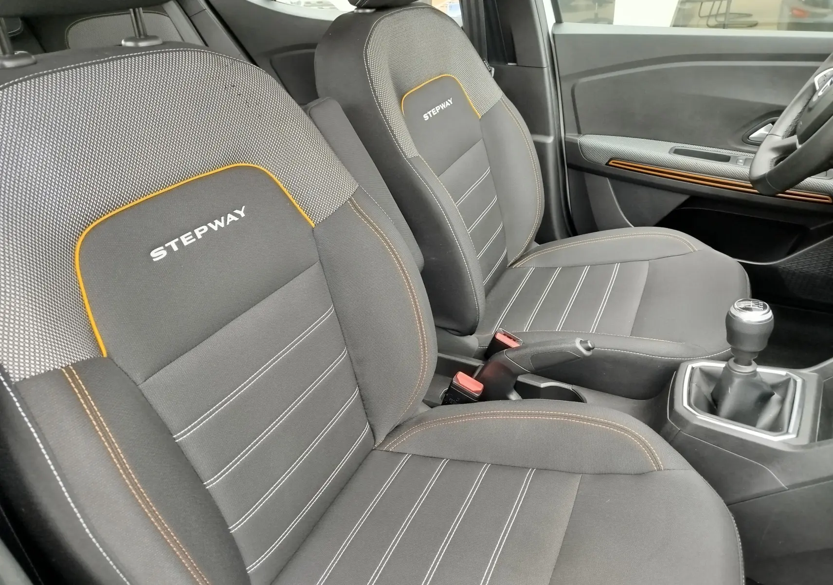 Vue intérieure côté conducteur de la Dacia Sandero Stepway 2021 avec sièges gris et surpiqûres orange, boîte manuelle visible.