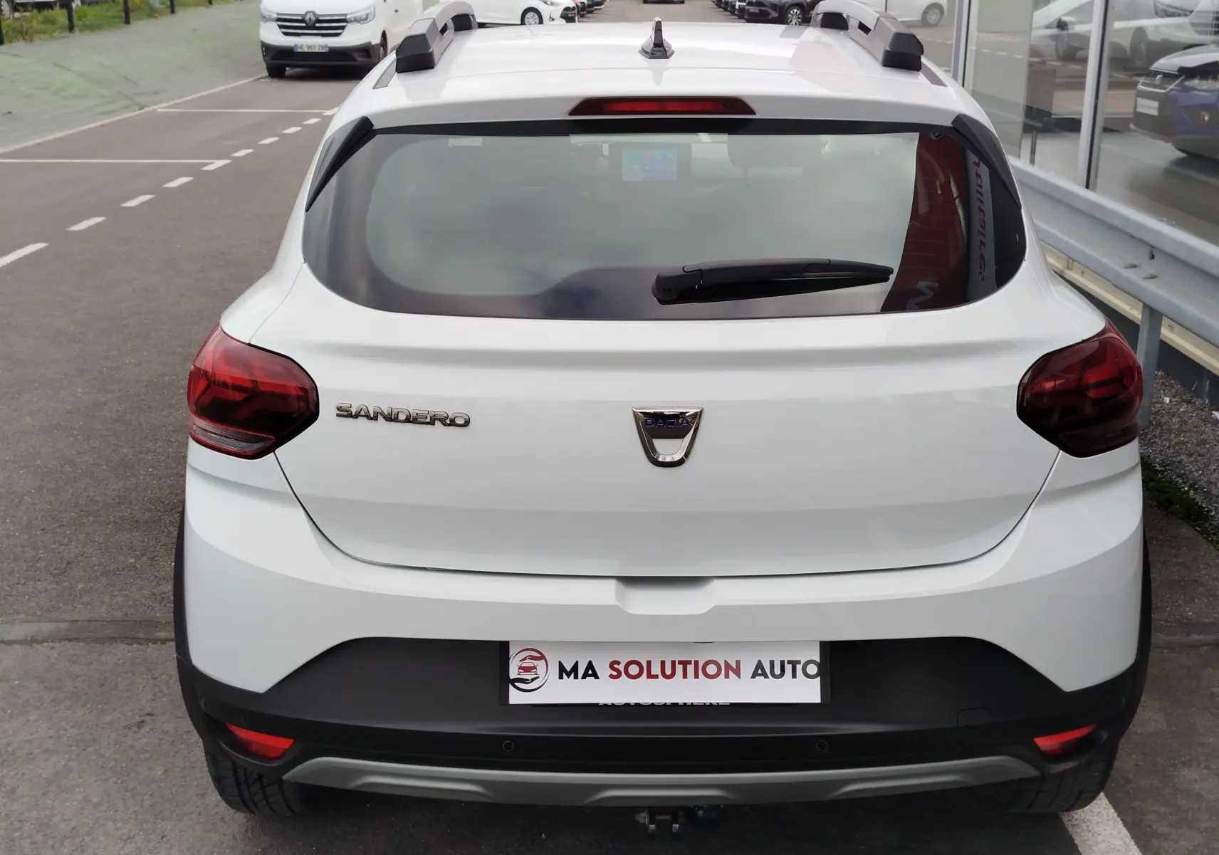 Vue arrière d'une Dacia Sandero Stepway blanche glacier avec feux arrière rouges et barres de toit noires.
