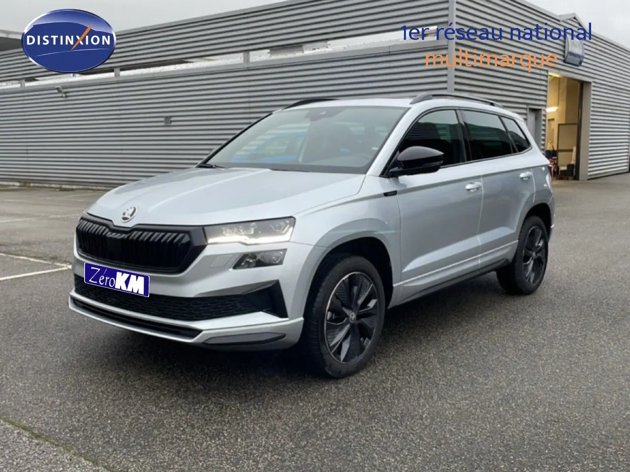 Vue 3/4 avant d'un Skoda Karoq gris argent métallisé avec jantes noires et calandre noire, stationné devant un bâtiment industriel.