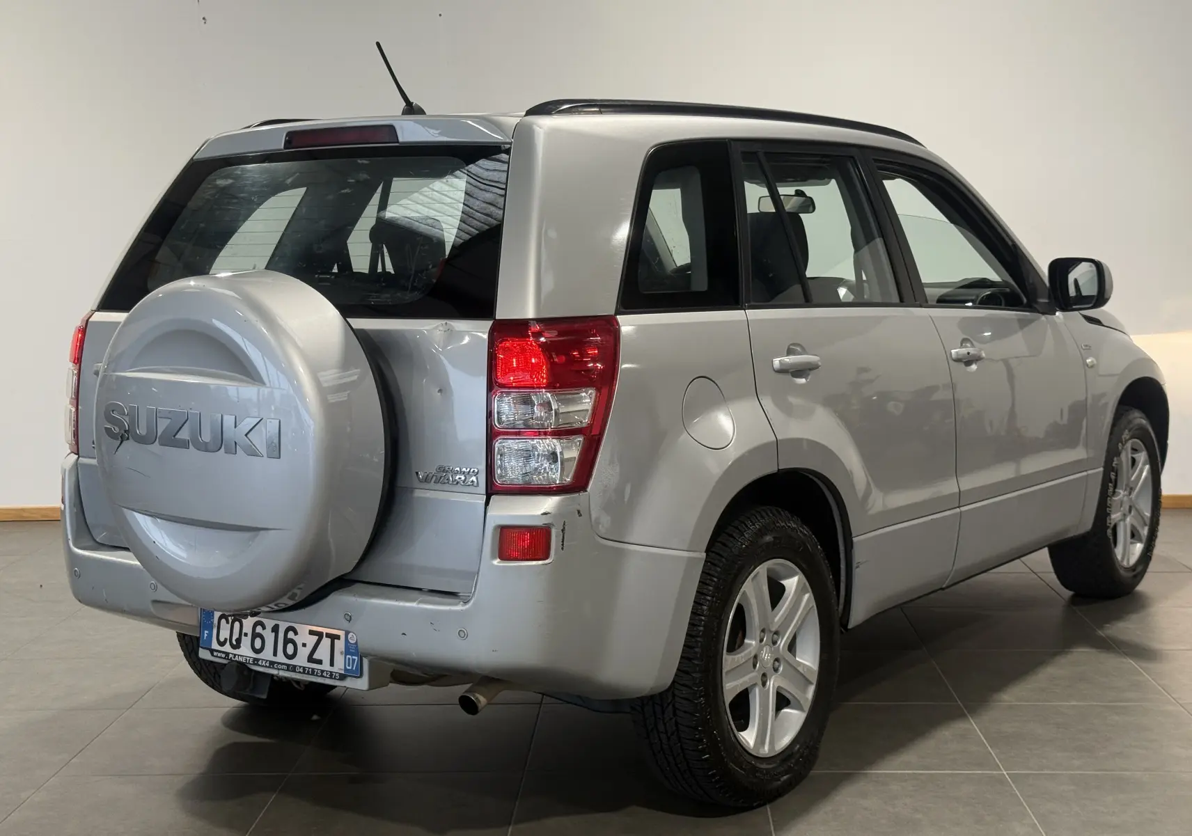Suzuki Grand Vitara gris 3/4 arrière droit avec roue de secours sur porte arrière, intérieur visible par les vitres.