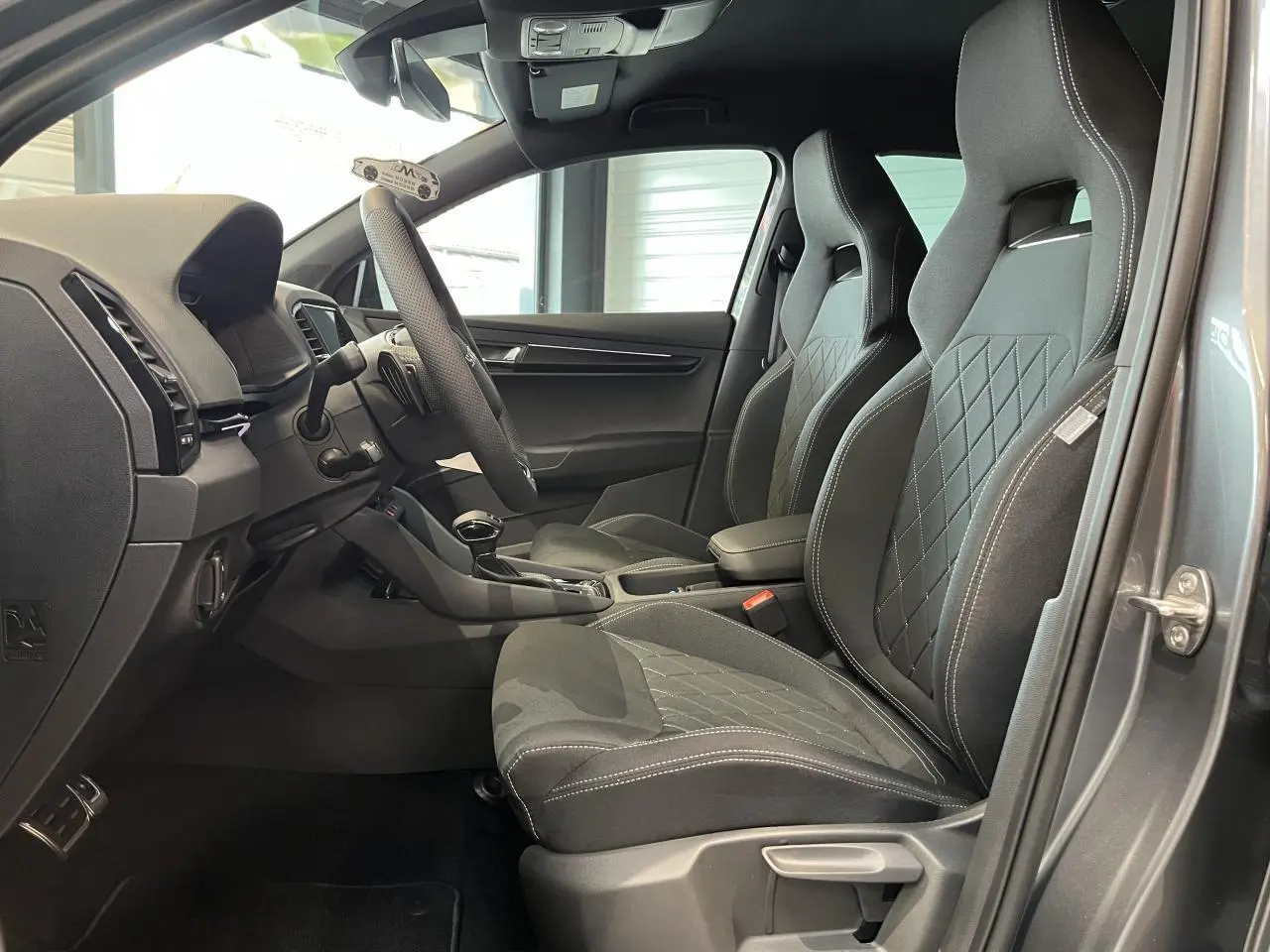 Vue intérieure côté gauche du Skoda Karoq gris 2025, mettant en valeur les sièges sport noirs avec surpiqûres blanches.