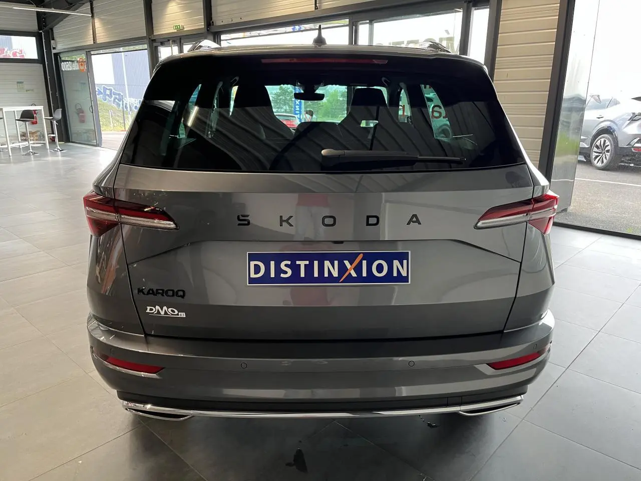 Vue arrière d'un SUV SKODA KAROQ gris métallisé exposé en showroom avec logo Distinxion sur le hayon.