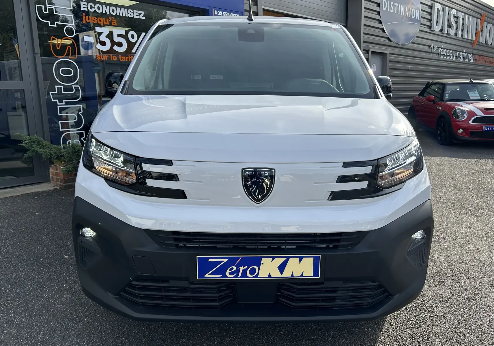 Vue de face d'un Peugeot Partner Fourgon blanc avec calandre noire et plaque Zéro KM, stationné devant un garage.