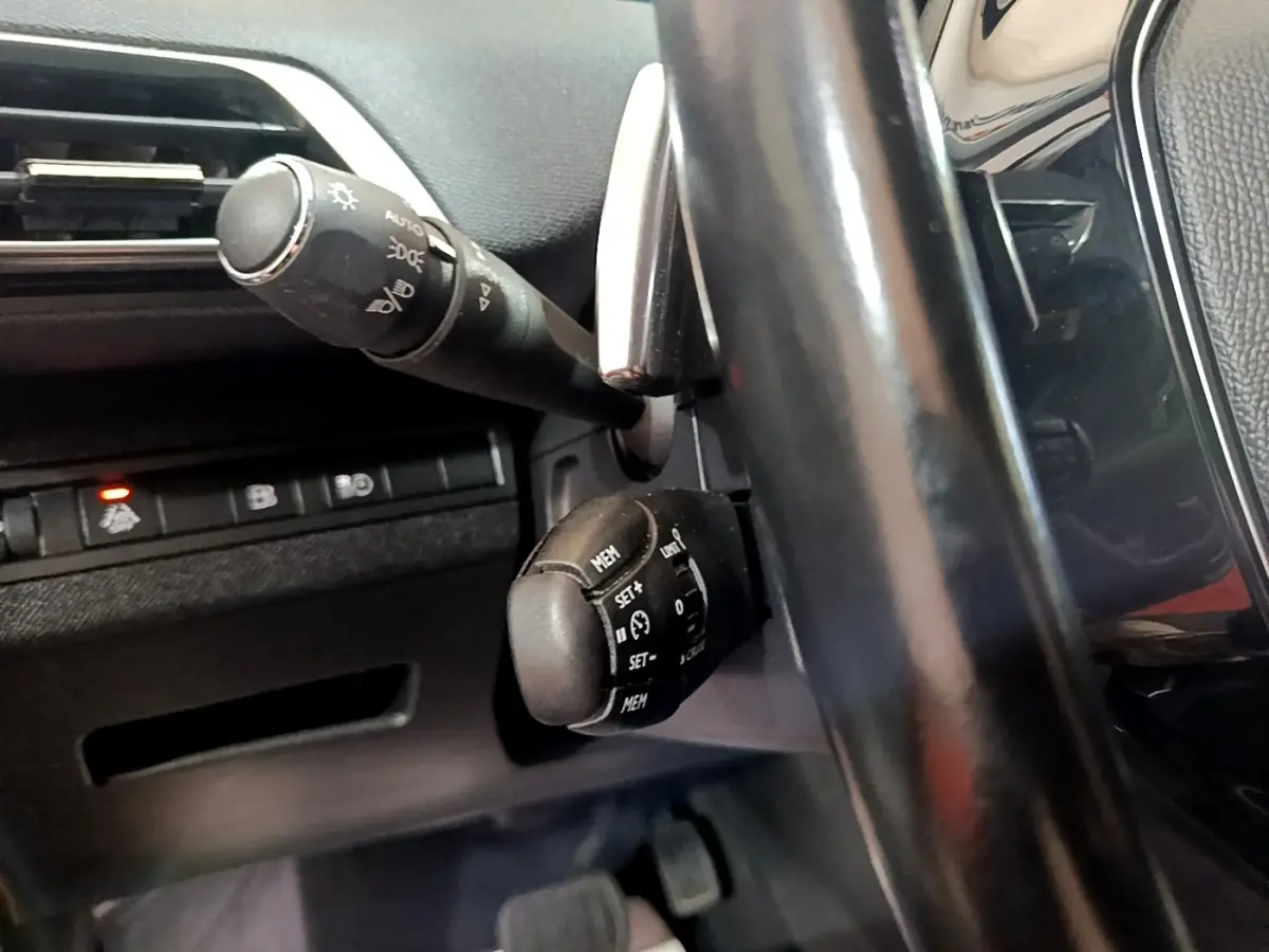 Gros plan sur la commande de régulateur de vitesse derrière le volant noir du Peugeot 3008 hybride blanc, côté gauche.