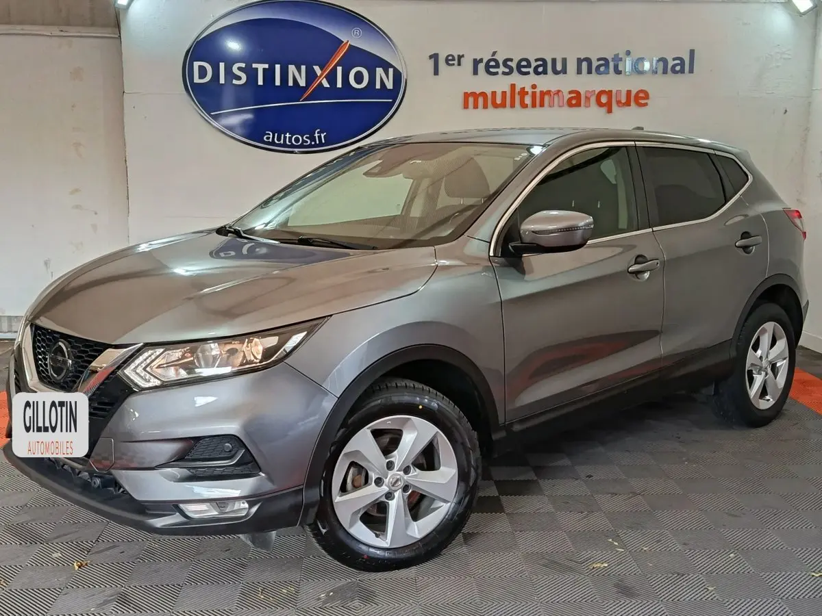 Nissan Qashqai gris clair vu de 3/4 avant droit, avec jantes alliage et phares allumés en intérieur showroom.