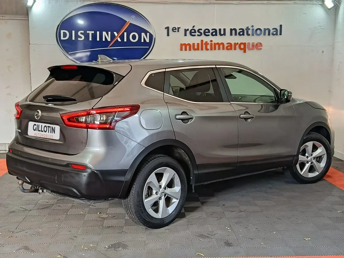 Nissan Qashqai gris clair vu de trois quarts arrière droit, avec feux arrière et jantes visibles en intérieur showroom.