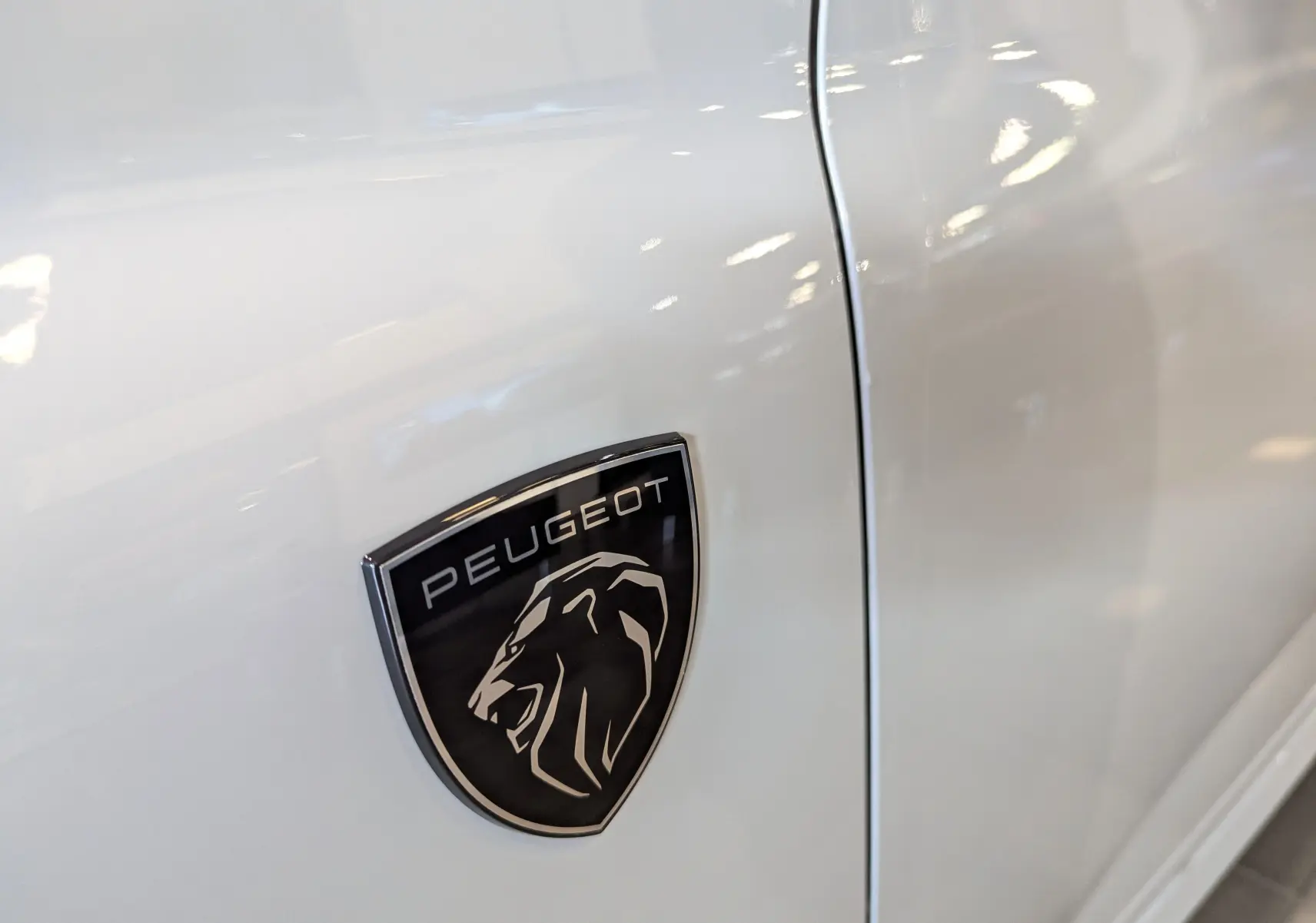 Gros plan sur l'écusson noir Peugeot avec lion argenté sur la porte blanche Okénite d'une 308 GT 2025.